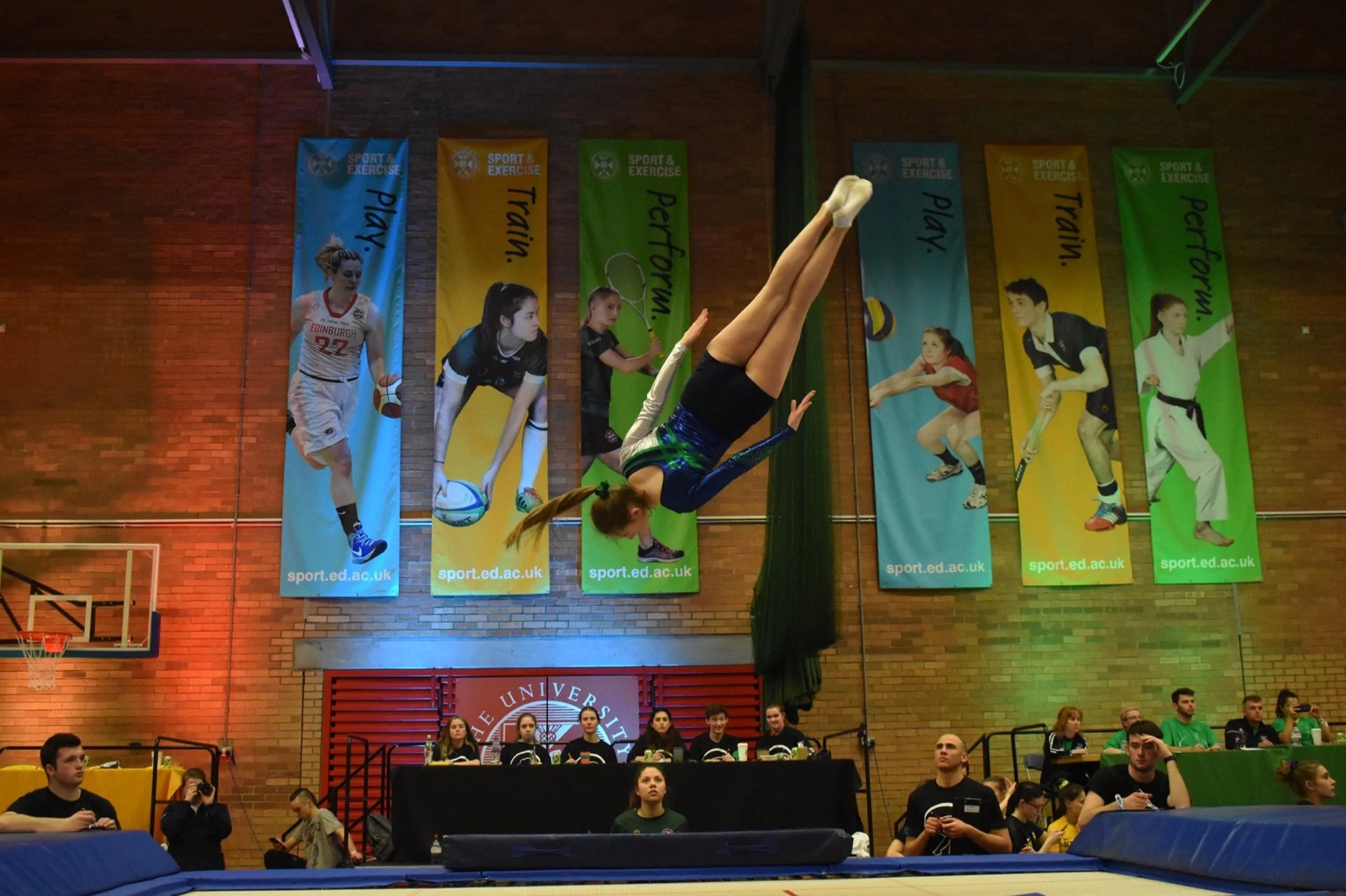 A picture of a female gymnast performing a straight somersault, updside-down in the air. She is wearing the EUTC green and blue leotard.